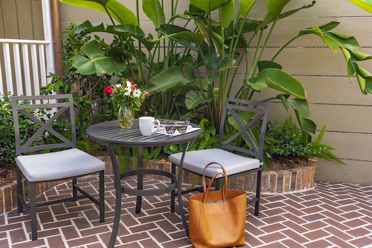 A patio scene with two chairs, a table, flowers, a mug, sunglasses, a magazine, and a tote bag, surrounded by lush green plants.