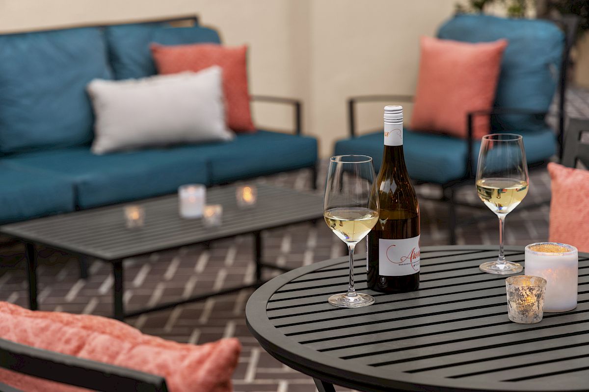 A cozy patio setting with a bottle of white wine, two glasses on a table, candles, and blue and orange cushions.